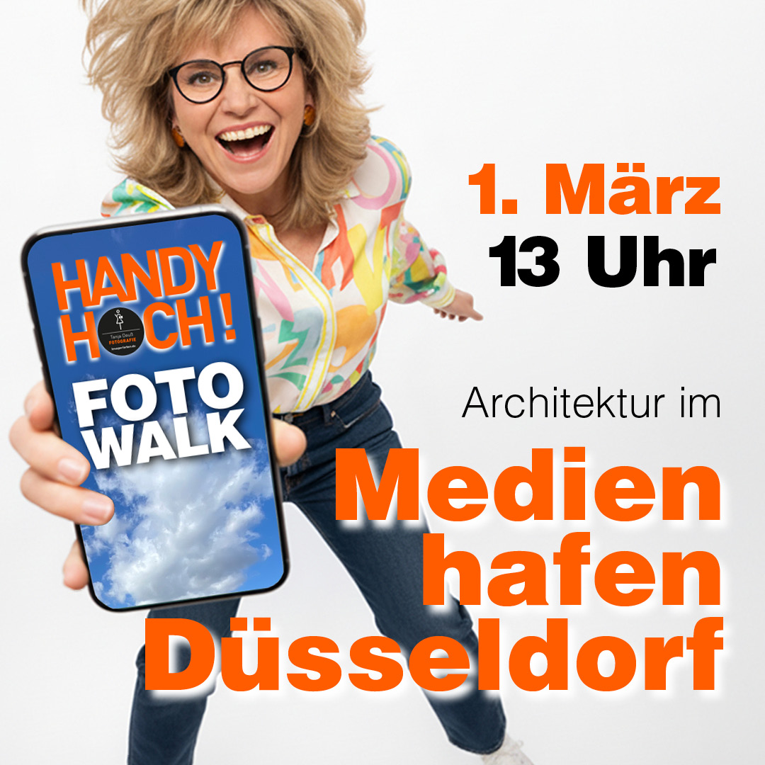 Frau hält iPhone in die Kamera. Auf dem Display steht: HANDY HOCH! Foto Walk Text im Bild: im Medienhafen Düsseldorf. 1. März 2023 um 13 Uhr.